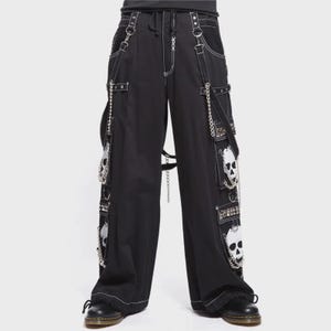 Puede incluir: Pantalones negros de pierna ancha con costuras blancas en contraste, múltiples bolsillos cargo con gráficos de calaveras blancas, cadenas metálicas plateadas, anillas en D y correas con tachuelas. Incluyen cintura con cordón y paneles de malla.