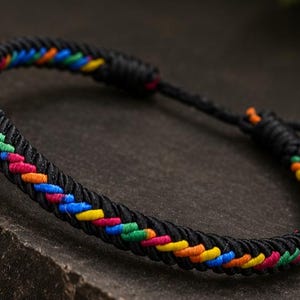 May include: A black cord bracelet featuring a braided pattern with vibrant multi-coloured accents in red, orange, yellow, green, and blue. The bracelet includes an adjustable sliding knot closure with two thin black cord ends finished with small knots.
