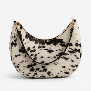 May include: A crescent-shaped shoulder bag featuring a white and black cow-print pattern with a textured hair-on-hide finish. The bag includes a thin black strap attached with gold-toned metal clasps and a top zipper closure along the curved edge.