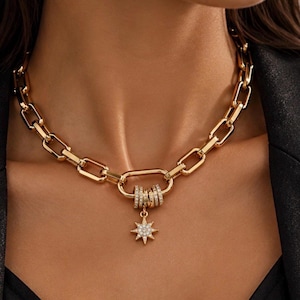 Puede incluir: Un collar de cadena en tono dorado con eslabones rectangulares gruesos entrelazados. Un gran cierre tipo mosquetón ovalado sirve como punto focal, adornado con varios anillos pequeños brillantes y un colgante en forma de estrella con cristales.