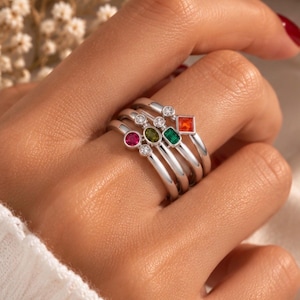 Puede incluir: Un conjunto de cuatro anillos de plata con varias piedras preciosas. Los anillos incluyen una piedra rosa redonda, una piedra verde oliva ovalada, una piedra verde esmeralda rectangular y una piedra naranja cuadrada, con pequeños detalles de piedras claras.