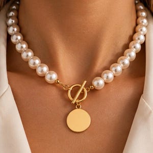Peut inclure: Un collier composé d'un rang de perles rondes de couleur crème. Le collier est fermé par un fermoir à bascule en métal doré situé à l'avant, agrémenté d'un pendentif circulaire doré suspendu au centre de l'anneau du fermoir.