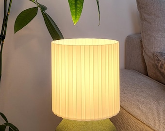 Peut inclure: Une lampe de table dotée d'un abat-jour cylindrique blanc à plis verticaux diffusant une lumière chaude, reposant sur une base arrondie vert olive texturée. La lampe est posée sur une surface en bois à côté d'un livre à couverture fleurie.