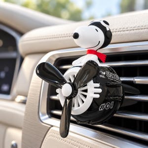 Pode incluir: Um ambientador de carro preto e branco em forma de avião vintage com uma figura do Snoopy a usar um chapéu de piloto e um cachecol vermelho. A hélice é preta e o lado do avião apresenta o texto Snoopy e o número 01 em branco.