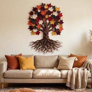May include: A large wall hanging featuring a tree with a twisted brown trunk and exposed roots, adorned with numerous fabric leaves in shades of red, orange, yellow, cream, and dark green, mounted on a plain off-white wall above a sofa.