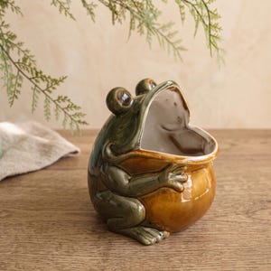 May include: A ceramic frog-shaped container with a glossy finish. The frog has a dark green back and head with a light brown, rounded belly. Its mouth is wide open, creating a hollow interior space. The eyes are prominent and dark.