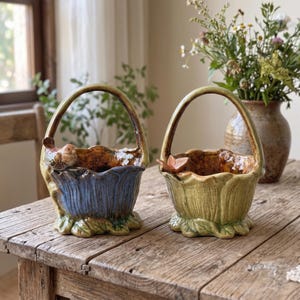 May include: Two ceramic baskets with arched handles and scalloped rims. The left basket is blue with a small brown bird perched on the edge. The right basket is green with a small brown butterfly on the rim. Both feature leaf-like bases.