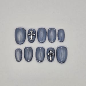 May include: A set of ten press-on nails in a glossy slate blue finish. Each nail features small silver-toned studs along the perimeter. Two of the nails are accented with a prominent, three-dimensional silver-toned cross charm centered on the surface.