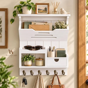 May include: A white wall-mounted wooden organizer featuring a top shelf, a mail slot, a middle shelf with a small drawer, and six bronze hooks. The unit holds a potted plant, sunglasses, pens, a notebook, a candle, and various hanging items.