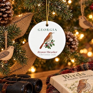 May include: A round white ceramic ornament featuring a brown thrasher bird perched on a holly branch. The text GEORGIA appears in dark green above the bird, while Brown Thrasher and Christmas 2026 are printed in dark red below the illustration.