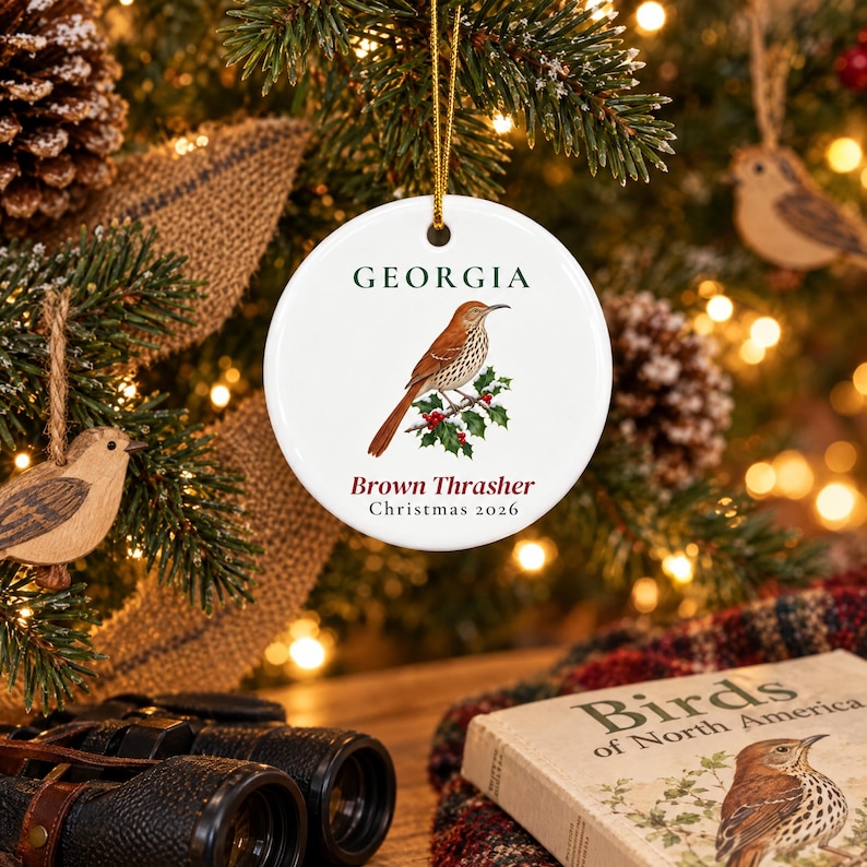 May include: A round white ceramic ornament featuring a brown thrasher bird perched on a holly branch. The text GEORGIA appears in dark green above the bird, while Brown Thrasher and Christmas 2026 are printed in dark red below the illustration.