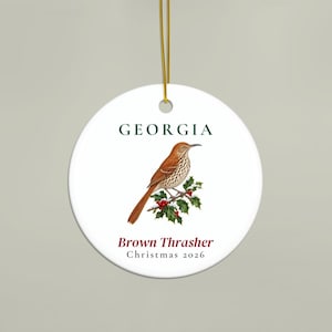 May include: A round white ceramic ornament featuring a brown thrasher bird perched on a holly branch with red berries. The text GEORGIA appears in dark green above the bird, with Brown Thrasher and Christmas 2026 printed in dark red and black below.
