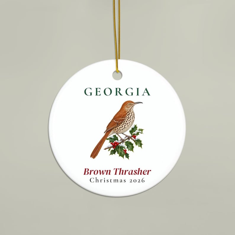 May include: A round white ceramic ornament featuring a brown thrasher bird perched on a holly branch with red berries. The text GEORGIA appears in dark green above the bird, with Brown Thrasher and Christmas 2026 printed in dark red and black below.