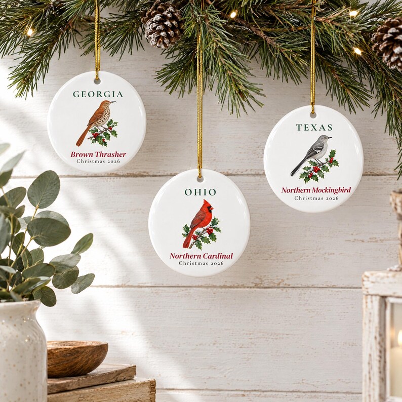 May include: Three round white ceramic ornaments hanging by gold strings. Each features a state name, a bird illustration, the bird's common name, and the text Christmas 2026. The birds are a Brown Thrasher for Georgia, a Northern Cardinal for Ohio, and a Northern Mockingbird for Texas.