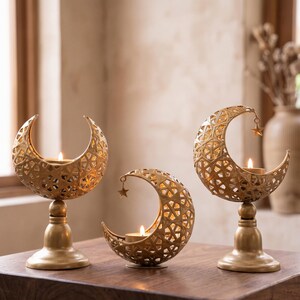 May include: Three gold-colored metal candle holders shaped like crescent moons with intricate geometric cutouts. Two holders stand on pedestals, while one sits directly on the surface. Each holds a small lit tea light candle.