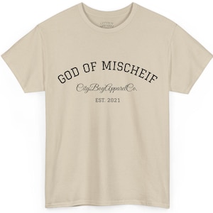 May include: A beige short-sleeved t-shirt featuring black text on the chest that reads GOD OF MISCHIEF in an arched font, followed by the script text CityBoyApparelCo. and the printed text EST. 2021 centred below the main graphic.