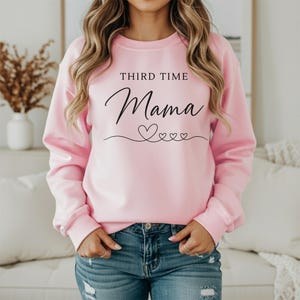 May include: A light pink long-sleeve sweatshirt featuring the black text "THIRD TIME Mama" printed on the chest, accompanied by a decorative line drawing of four small hearts connected by a continuous flowing line underneath the lettering.