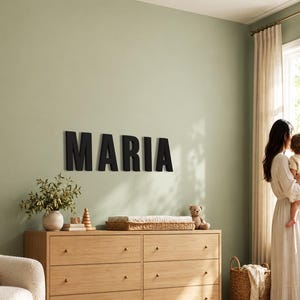 Op de afbeelding: Grote zwarte hoofdletters die de naam MARIA vormen, zijn bevestigd aan een saliegroene muur boven een lichte houten ladekast met zes lades. Op de ladekast staan een kleine plant in een pot, een houten stapelspeeltje en een gevlochten dienblad.