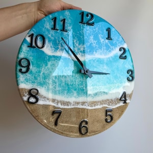 Puede incluir: Un reloj de pared redondo con un diseño de resina brillante que muestra una playa de arena y olas turquesas con espuma blanca. La esfera tiene números negros en relieve del 1 al 12 y manecillas negras para horas, minutos y segundos.