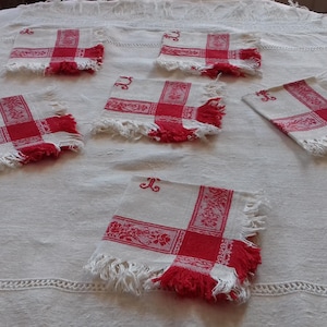 Op de afbeelding: Een set van vijf vierkante, gebroken witte linnen servetten met een rood kruispatroon, bloemmotieven en franjes. Elk servet heeft een klein rood geborduurd monogram in de bovenhoek, gerangschikt op een bijpassend gebroken wit tafelkleed.
