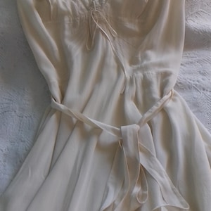 Può includere: Un abito color crema caratterizzato da uno scollo a V con delicate rifiniture in pizzo, maniche corte a volant e una cintura in tessuto abbinata in vita. Il capo presenta una silhouette morbida e fluida con sottili cuciture verticali.