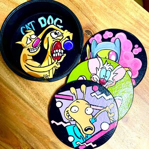Pode incluir: Um conjunto de bases para copos pintadas à mão com personagens de desenhos animados. Uma base para copos tem o texto "CAT DOG" com os personagens CatDog. Outras bases para copos apresentam Pinky e Cérebro, e Rocko. As bases para copos são pretas com desenhos coloridos.