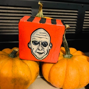 May include: Orange pumpkins with a painted wooden block featuring a face with a bald head and dark eyes. The block is painted orange with black stripes and is resting on top of the pumpkins. The pumpkins are a bright orange color.