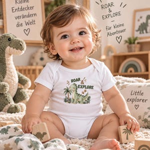 Könnte beinhalten: Ein weißer kurzärmeliger Babybody mit einem grünen Dinosaurier-Motiv, Palmen und dem Text ROAR & EXPLORE. Das Design enthält kleine braune Dinosaurier-Fußabdrücke und ist mittig auf der Brust des Kleidungsstücks platziert.