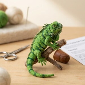 Puede incluir: Una figura de iguana verde hecha con fieltro de aguja posada sobre una pequeña rama marrón. La iguana presenta piel texturizada, una cresta dorsal espinosa y rayas de color verde oscuro a lo largo del cuerpo y la larga cola de lana.