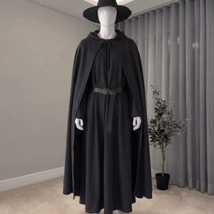 以下が含まれることがあります： 黒いフード付きマントと長袖の黒いドレスのセット。ウエストは長方形のバックルが付いた黒いベルトで締められています。つばの広い黒いとんがり帽子を合わせた衣装が、白いマネキンに着せられています。
