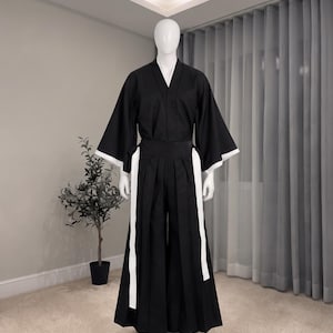 Könnte beinhalten: Ein schwarzes Oberteil im Kimono-Stil mit weiten Ärmeln und weißen Manschetten, kombiniert mit einer schwarzen, plissierten Hakama-Hose mit langen weißen seitlichen Bindebändern. Das Outfit wird auf einer weißen Schaufensterpuppe präsentiert.