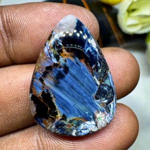 Può includere: Una pietra preziosa di pietersite a forma di goccia con una superficie lucida e motivi vorticosi di fibre chatoyant blu profondo, nere e marrone dorato. La pietra presenta un taglio cabochon liscio con una finitura lucida.
