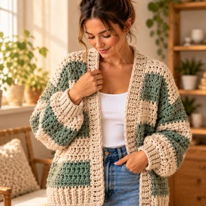 May include: A chunky crochet cardigan featuring a color-blocked pattern of alternating cream and sage green rectangular panels. The garment has long sleeves with ribbed cuffs, a ribbed hem, and an open front design with a thick ribbed border.