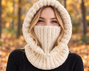 Könnte beinhalten: Eine cremefarbene, grob gestrickte Sturmhaube mit einer dicken, gerippten Textur, die Kopf, Hals und das untere Gesicht bedeckt. Die Kopfbedeckung besteht aus weichem, dickem Garn und bietet vollständige Abdeckung mit einer weiten Öffnung.