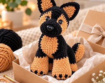 Könnte beinhalten: Ein gehäkeltes Schäferhund-Stofftier aus schwarzem und beigem Garn. Das Kuscheltier hat große spitze Ohren, eine abgerundete Schnauze mit einer schwarzen Kunststoffnase und dunkle glänzende Augen und sitzt aufrecht in einer Pappschachtel.