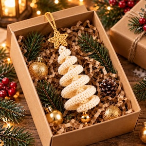 May include: A cream-colored spiral crochet Christmas tree ornament topped with a matching star, presented in a brown cardboard gift box filled with shredded paper, green pine sprigs, small gold baubles, and a single brown pinecone.
