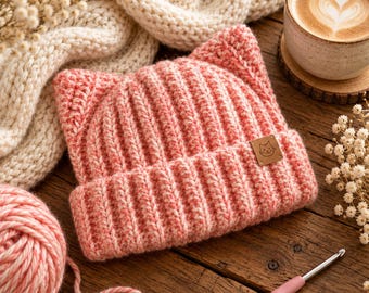 Könnte beinhalten: Eine gehäkelte rosa Mütze mit zwei spitzen Katzenohren oben. Die Mütze hat eine gerippte Textur und ein kleines quadratisches braunes Lederetikett auf der Vorderseite mit einer eingravierten Strichzeichnung eines Katzengesichts.