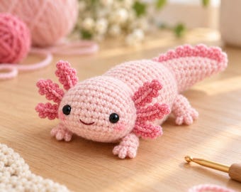 Könnte beinhalten: Ein hellrosa gehäkeltes Axolotl-Plüschtier mit strukturiertem Körper, langem Schwanz, kleinen Gliedmaßen und markanten äußeren Kiemen an den Kopfseiten. Das Wesen hat zwei kleine schwarze Perlenaugen und ein aufgesticktes Lächeln.