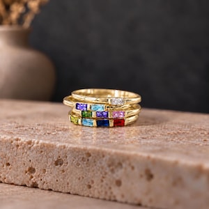 May include: A stack of three gold rings, each featuring a row of rectangular baguette-cut gemstones in various colors including red, blue, green, purple, pink, and clear, set horizontally along the thin, polished gold bands.