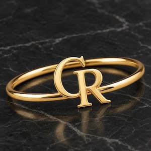 May include: A polished gold-tone bangle bracelet featuring a prominent, centred capital letter C and capital letter R monogram. The metallic surface of the circular band and the letters reflects light, creating a smooth, shiny, and reflective finish.