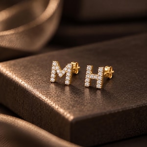May include: A pair of gold stud earrings shaped like the letters M and H, each featuring a pavé setting of small, clear, sparkling stones. The earrings have a polished gold finish and include standard post backs for secure attachment.