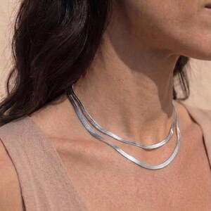 Könnte beinhalten: Eine zweilagige silberne Schlangenkette mit flachem, flexiblem Glieddesign, das am Hals anliegt. Die metallisch silberne Oberfläche reflektiert das Licht und verleiht diesem geschichteten Schmuckstück ein glattes, durchgehendes Erscheinungsbild.