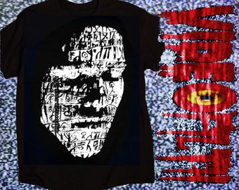 May include: A black short-sleeve t-shirt featuring a white graphic of a face composed of various symbols and text. To the right, the word "VIDEODROME" is printed in a red, dripping font with a yellow eye graphic centered in the letter O.