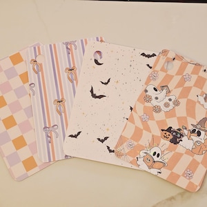 May include: Four rectangular cards with rounded corners and a hole punch in the top corner. Designs include a lavender and orange checkerboard, vertical stripes with orange bows, a cream background with black bats, and a wavy peach checkerboard with ghosts.
