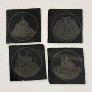 May include: A set of four square black slate coasters, each featuring a circular engraved design of a different historic castle or landmark. The dark stone surfaces have a natural, textured appearance with detailed architectural illustrations.