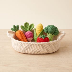 May include: A round, cream-colored crocheted basket with two side handles containing a collection of crocheted play vegetables, including an orange carrot, a pink radish with green leaves, a yellow corn cob, a red tomato, and a green broccoli.