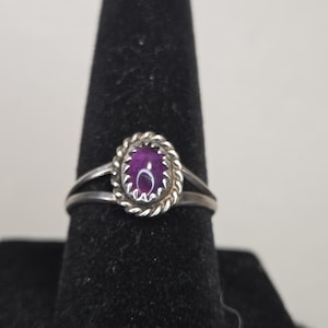 May include: A silver ring featuring an oval-shaped purple gemstone set in a twisted rope-style bezel. The band of the ring splits into two separate silver shanks as it connects to the central setting, creating a simple and clean design.