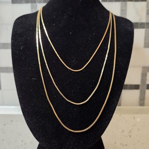 May include: A set of three gold-toned chain necklaces displayed in a layered arrangement. Each necklace features a flat, flexible herringbone-style link design that reflects light, creating a smooth, continuous metallic appearance across the chains.