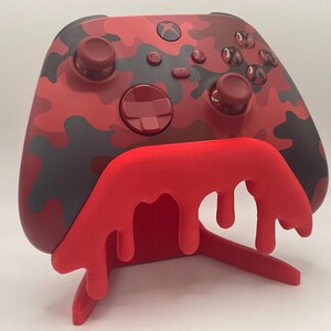 Könnte beinhalten: Ein rot-schwarz camouflierter Videospiel-Controller auf einem leuchtend roten Ständer mit einem dekorativen, tropfenden Flüssigkeitsdesign. Der Controller verfügt über zwei Analogsticks, ein Steuerkreuz und verschiedene Tasten mit dem Xbox-Logo.