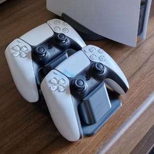 Könnte beinhalten: Zwei weiß-schwarze kabellose Gamecontroller in einer dunkelgrauen Ladestation aus Kunststoff. Die Controller verfügen über zwei Analogsticks, ein Steuerkreuz und vier Aktionstasten. Eine weiße Spielkonsole ist im Hintergrund teilweise zu sehen.